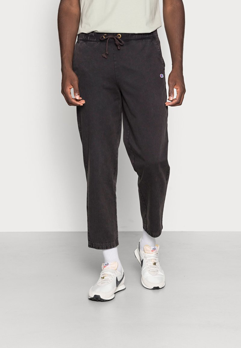 Pantalones de mezcla de algodón negros con cintura elástica y cordón. Piernas ajustadas y un pequeño logotipo en la cadera derecha. Llevados con zapatillas blancas.