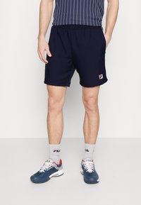 Shorts de sport navy avec taille élastique, fabriqués dans un matériau léger. Portés avec des chaussettes grises et des baskets Fila navy/rouge. Rayures horizontales sur le t-shirt.