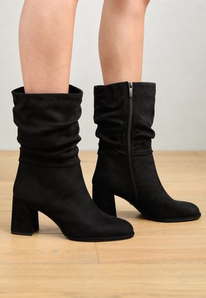 Bottes mi-mollet en daim noir avec talons carrés et fermeture éclair latérale portées sur jambes nues sur un fond de sol en bois.