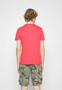 Persona joven con cabello rizado que lleva una camiseta roja lisa y pantalones cortos de camuflaje verde, de espaldas con un fondo blanco liso.