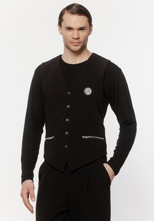 Mężczyzna ubrany w czarną zapinaną na guziki kamizelkę z kieszeniami na zamek, na czarnej koszuli z długim rękawem, stojący z jedną ręką w kieszeni.