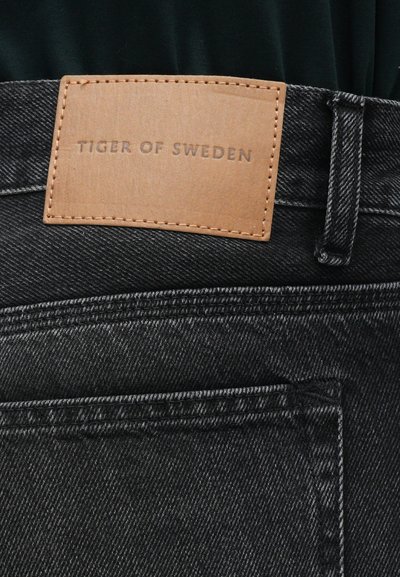 Jeans din denim negru cu o etichetă din piele inscripționată "TIGER OF SWEDEN." Prezintă cusături elaborate și un design clasic cu cinci buzunare.