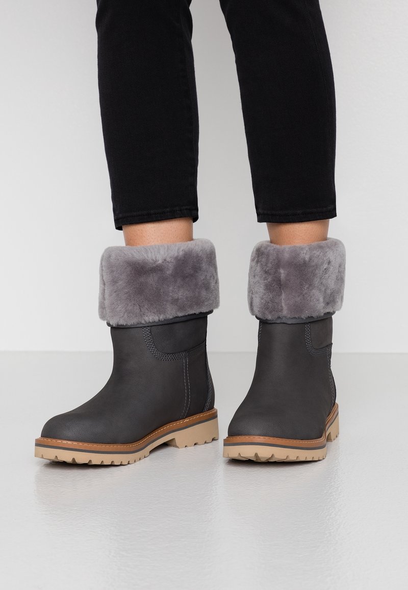 Bottes noires à hauteur de cheville avec des semelles épaisses beige et des poignets en fourrure grise pliée, portées avec un pantalon noir sur un sol blanc.