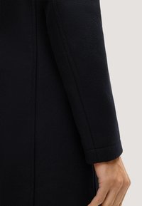 Manica di un cappotto nero in tessuto testurizzato, con design sartoriale e orlo pulito. Il materiale sembra robusto, migliorando la durata e la vestibilità.