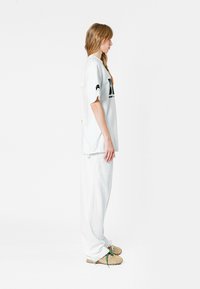 Camiseta blanca de gran tamaño con mangas cortas, gráfico negro y pantalones blancos holgados. El modelo lleva zapatillas beige con cordones verdes.