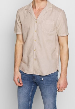 Mand iført en beige kortærmet skjorte med knapper og brystlomme samt blå jeans, stående mod en ensartet lys baggrund.