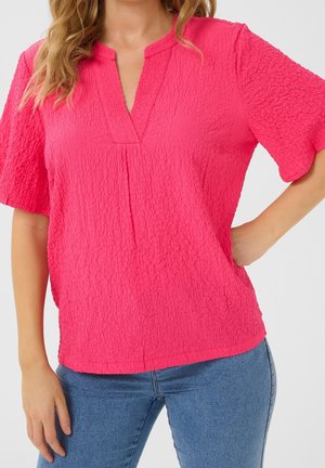 Vrouw draagt een felroze blouse met korte mouwen en een V-hals, gecombineerd met een blauwe spijkerbroek, staand met één hand op de heup tegen een effen achtergrond.