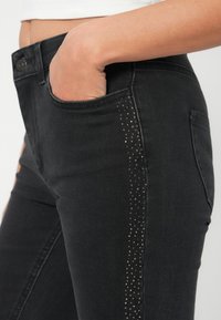 Vaqueros de mezclilla negra de corte slim, con un acento lateral de pequeños clavos metálicos a lo largo de la costura izquierda y un bolsillo frontal.