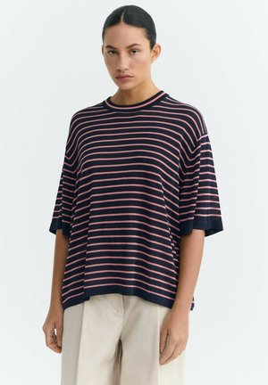 Junge Frau mit dunklem Haar zu einem niedrigen Dutt gebunden, trägt ein locker sitzendes marineblaues Hemd mit rosa horizontalen Streifen und beige Hosen vor einem einfarbigen Hintergrund.