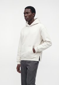 Homme debout portant un sweat à capuche blanc avec poche avant et pantalon gris sur un fond blanc uni.