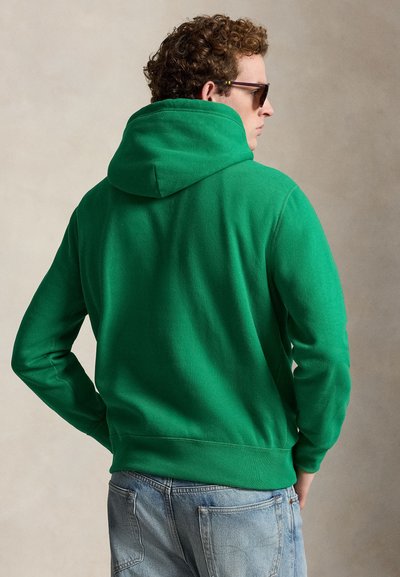 Homme aux cheveux bouclés portant des lunettes de soleil foncées, un sweat à capuche vert et un jean bleu clair, tourné de dos sur un fond uni.
