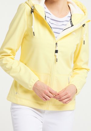 Person trägt eine hellgelbe Kapuzenjacke mit Reißverschluss über einem weiß-navyblau gestreiften Hemd, weiße Hose, Hände vor dem Körper verschränkt.