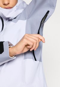 Lahka, vodoodporna jakna v beli in sivi barvi z teksturiranim materialom, opremljena z zadrgo na rokavu in velcro zapiranjem na zapestju.