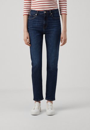 Personne portant un jean bleu foncé à jambes droites, des baskets blanches et un t-shirt à manches longues rayé rouge et blanc, debout devant un fond uni.