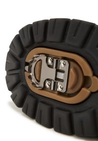 Suela de goma negra con profundos relieves y un mecanismo de hebilla metálica en un acabado bronce; resalta la durabilidad y el diseño funcional.