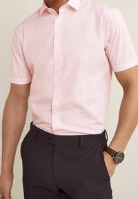 Homme portant une chemise à manches courtes rose clair boutonnée, rentrée dans un pantalon sombre, avec une montre-bracelet au poignet gauche.