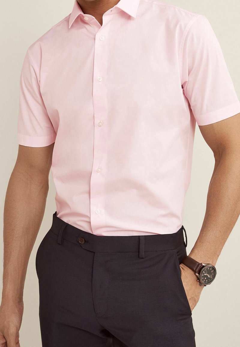 Homme portant une chemise à manches courtes rose clair boutonnée, rentrée dans un pantalon sombre, avec une montre-bracelet au poignet gauche.