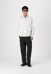 Weißes Langarmshirt mit vertikalen marineblauen Streifen, schwarze, locker sitzende Hose und schwarze klobige Schuhe; der Träger trägt Sonnenbrillen.