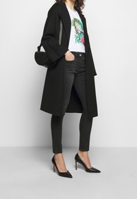 Black coat, white t-shirt with colorful floral print, black skinny jeans, and black pointed heels. Features a textured shoulder bag.