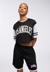 Top cropped in rete nera con la scritta "LOS ANGELES" in bianco, abbinato a pantaloni corti neri con bordo e logo bianchi. Entrambi realizzati in tessuto traspirante.