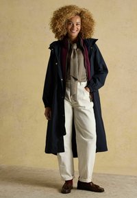 Palton marin lung peste un strat interior cărămiziu, bluză cu carouri, pantaloni albi cu croială largă și pantofi maronii cu accente roșii. Fundal texturat.