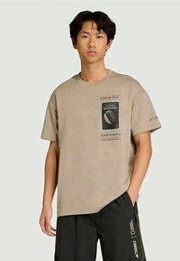 Fiatal férfi bézs National Geographic pólót visel, amelyen a "Nézz fel a Földről az űrbe" felirat található, fekete logós nadrággal, egy sima háttér előtt áll.