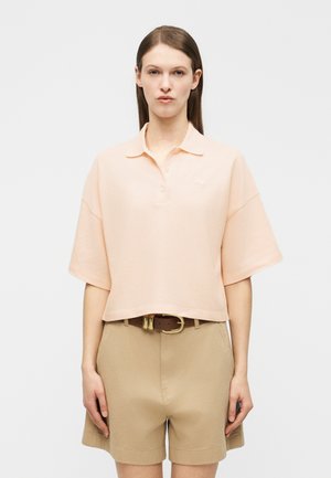 Žena sa ravnom smeđom kosom nosi blijedo breskvastu široku polo majicu koja je izvana preko bež kratkih hlača visokog struka s smeđim remenom.