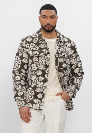 Man die een bruin-witte jas met bloemenpatroon draagt over een beige shirt en witte broek, staand met één hand in de zak.