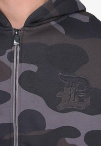Nærbillede af en grå og sort camouflagemønstret zip-hoodie med et broderet gotisk bogstav "D" emblem på brystet.