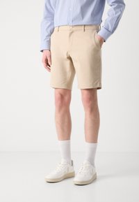 Beige shorts med en skreddersydd design og sidelommer, kombinert med hvite sneakers og hvite sokker i ankellengde. Blåstripete skjorte synlig.