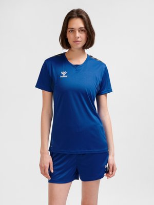 Vrouw staand, gekleed in een blauw hummel sportt-shirt en bijpassende short tegen een effen lichte achtergrond.
