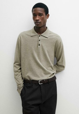 Jonge man draagt een lichtbeige poloshirt met lange mouwen, ingestopt in een zwarte broek met een bruine gevlochten riem, staand tegen een effen witte muur.