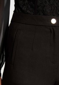 Pantalon noir avec une coupe ajustée, texture lisse, deux poches latérales et une fermeture à bouton argenté visible à la taille.