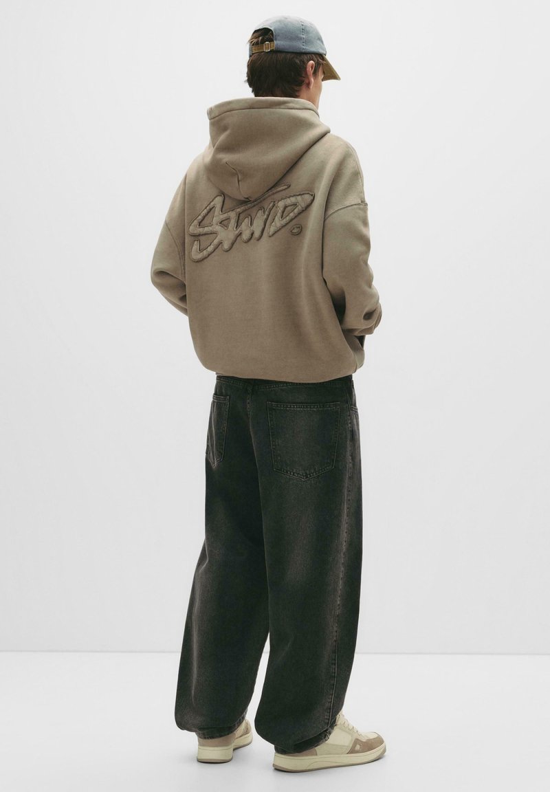 Sweatshirt i taupe med et tekstureret logo på ryggen, kombineret med løstsiddende mørk denim jeans og beige sneakers.