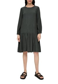 Dunkelgrünes Kleid mit langen Ärmeln, strukturiertem Stoff, rundem Ausschnitt und mehrlagigem Rüschensaum. Kombiniert mit schwarzen Sandalen.