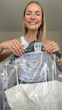 Bela obleka v plastični embalaži, ki jo drži oseba. Obleka ima brezramen kroj z nabranim krilom in oznako z informacijami o blagovni znamki.