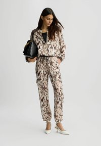 Beskåret jakke og joggers i beige med et brunt leopardprint. Tilbehør inkluderer en sort håndtaske og hvide spidse hæle.