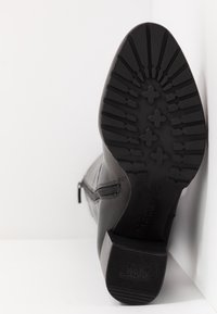 Botte en cuir noir avec semelle en caoutchouc à motif et talon bloc, fermeture éclair latérale visible, positionnée semelle vers le haut contre un coin de mur et de sol blancs.