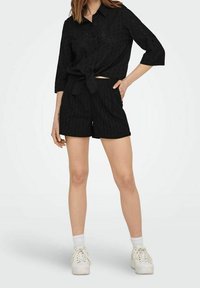 Camicia nera con bottoni, con nodo frontale, abbinata a pantaloni corti neri. Entrambi presentano un motivo testurizzato. Indossato con sneakers bianche e calze alla caviglia.