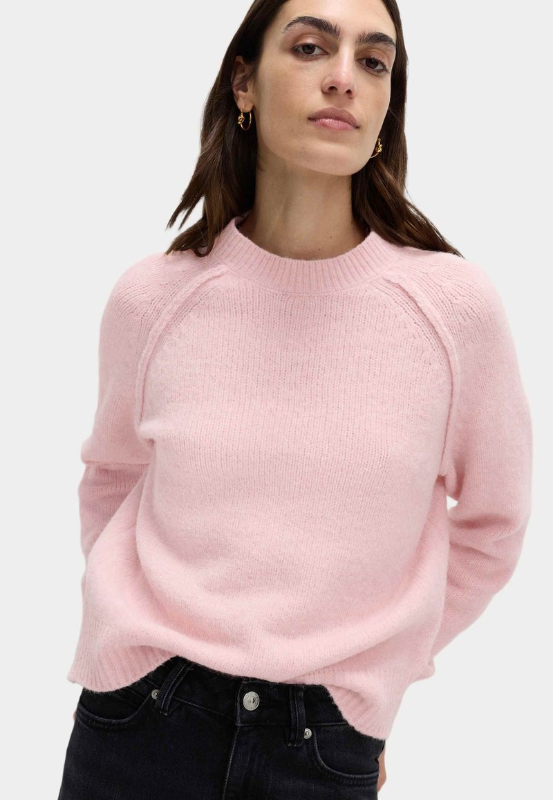 Maglione lavorato a maglia di un delicato rosa chiaro, con scollatura a costine e maniche raglan. Texture morbida abbinata a jeans scuri. Nessun motivo o accentuazione visibile.