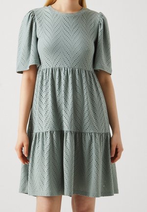 Femme portant une robe gris clair avec des motifs chevrons en œillets, des manches courtes bouffantes et une jupe à volants froncée.