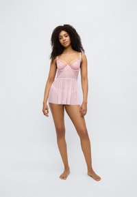 Σετ ροζ διάφανης εσωρούχου με απαλό, υφασμάτινο ύφασμα. Διαθέτει δαντελένιες λεπτομέρειες και ρυθμιζόμενους ιμάντες, σχεδιασμένο με εφαρμοστό μπούστο και ρέοντα φούστα.