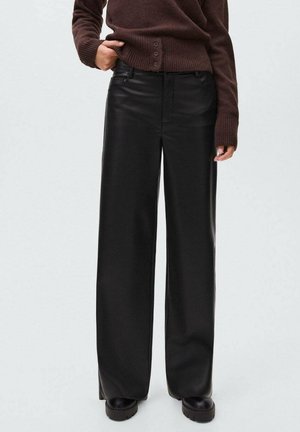 Personne portant un pantalon en cuir noir à taille haute et jambes larges, avec des chaussures à plateforme noires et un pull en tricot marron, se tenant avec une main dans la poche.
