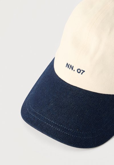 Casquette de baseball avec une calotte crème clair et une visière bleu marine foncé. Texte "NN. 07" brodé à l'avant, avec une texture de tissu lisse.
