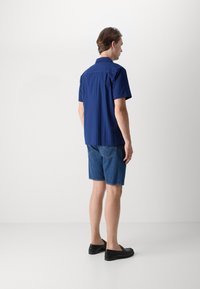 Modrá košeľa s krátkým rukávom a vertikálnymi pruhmi, v kombinácii s modrými riflovými šortkami. Čierne slip-on topánky dotvárajú vzhľad. Jednoduchý dizajn a pohodlný strih.