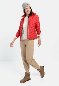 Rød quiltet jakke med hætte, beige bukser og brune ankelstøvler. Grå strikket hue. Model poserer mod en lys baggrund.