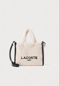 Bolso LACOSTE en color crema, con un exterior suave y texturizado, detalles en negro, dos asas cortas y una correa de hombro desmontable.