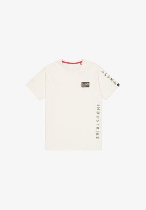 Krem majica kratkih rukava s crvenim izrezom. Ima pravokutnu kamuflažnu zakrpu na prsima i natpis "ALPHA INDUSTRIES" otisnut na strani.