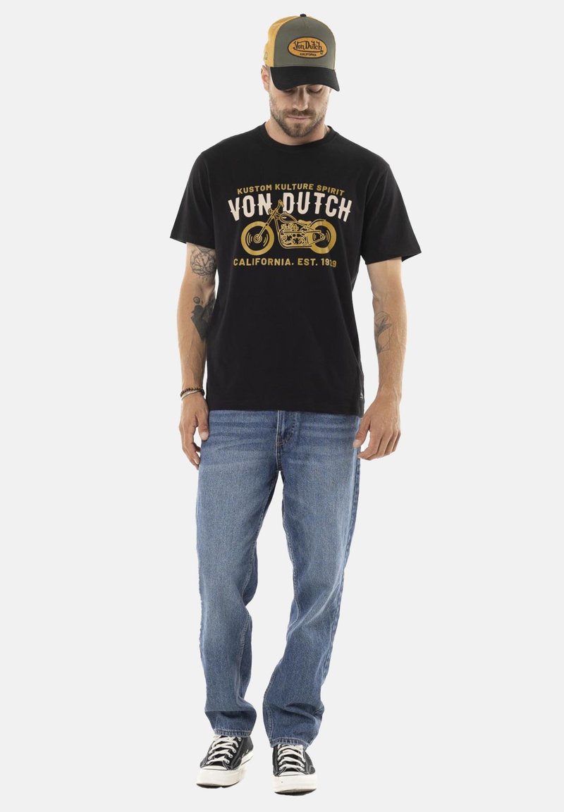 Camiseta de algodón negra con estampado gráfico dorado que presenta una motocicleta y texto. Combinada con vaqueros azules y zapatillas negras, rematada con una gorra negra y marrón.