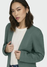 Groene blazer met een visgraatpatroon, gestructureerde schouders, open voorkant en een losse pasvorm, gecombineerd met een licht gestructureerde trui eronder.